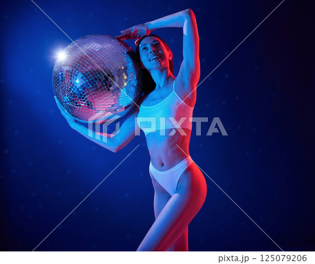 With shiny mirrored ball. Young woman in underwear is in the studio with neon lights With shiny mirrored ball. Young woman in underwear is in the studio with neon lights 125079206