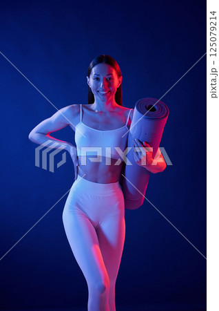 Front view. Holding the mat. Young woman in sportive clothes is in the studio with neon lights Front view. Holding the mat. Young woman in sportive clothes is in the studio with neon lights 125079214