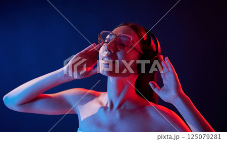 Enjoying music by using headphones. Portrait of young woman that is indoors in neon lighting Enjoying music by using headphones. Portrait of young woman that is indoors in neon lighting 125079281