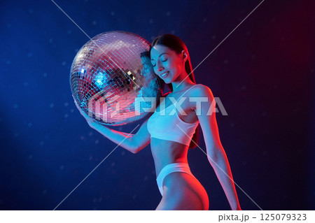 Party time. Holding mirrored ball. Young woman in underwear is in the studio with neon lights 125079323