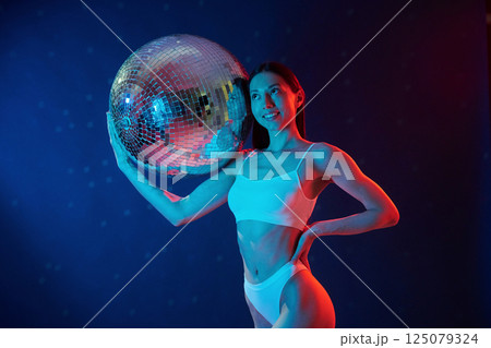 Party time. Holding mirrored ball. Young woman in underwear is in the studio with neon lights 125079324