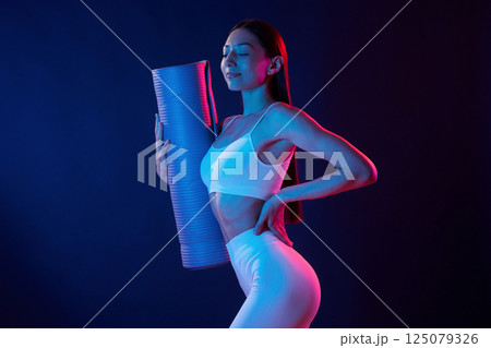 Young woman in sportive clothes is in the studio with neon lights. Holding mat 125079326