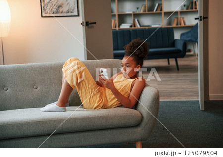 Surfing the internet by using smartphone. Cute black girl in casual clothes is at home at daytime 125079504