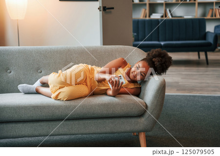 Surfing the internet by using smartphone. Cute black girl in casual clothes is at home at daytime Surfing the internet by using smartphone. Cute black girl in casual clothes is at home at daytime 125079505