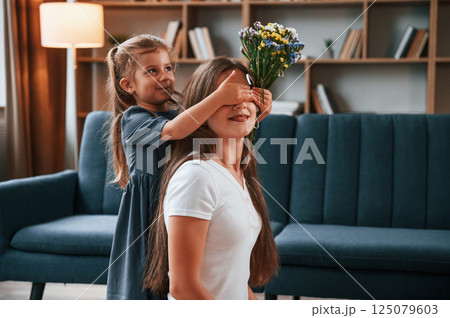 Girl is giving beautiful flowers to a woman. Young mother with her daughter is together at apartment 125079603