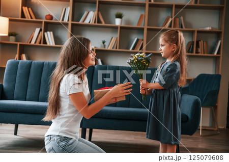 Girl is giving beautiful flowers to a woman. Young mother with her daughter is together at apartment 125079608