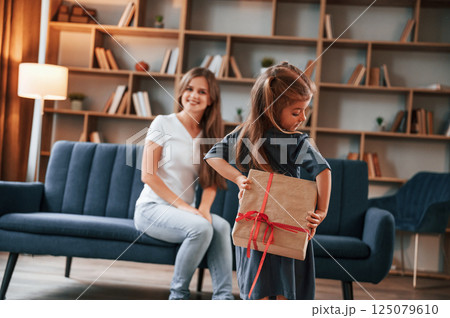Girl holding gift box behind her. Happy holidays. Young mother with her daughter is together at apartment Girl holding gift box behind her. Happy holidays. Young mother with her daughter is together at apartment 125079610