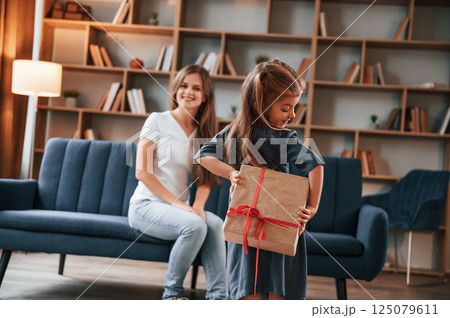 Girl holding gift box behind her. Happy holidays. Young mother with her daughter is together at apartment 125079611