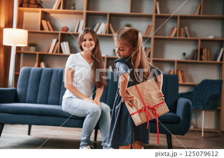 Girl holding gift box behind her. Happy holidays. Young mother with her daughter is together at apartment 125079612