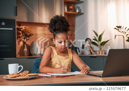 Sitting by the table with laptop and paper for drawing. Cute black girl in casual clothes is at home at daytime Sitting by the table with laptop and paper for drawing. Cute black girl in casual clothes is at home at daytime 125079677