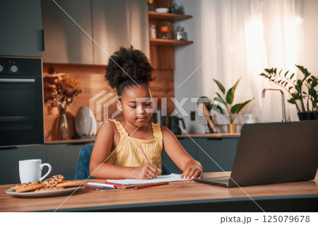 Sitting by the table with laptop and paper for drawing. Cute black girl in casual clothes is at home at daytime Sitting by the table with laptop and paper for drawing. Cute black girl in casual clothes is at home at daytime 125079678