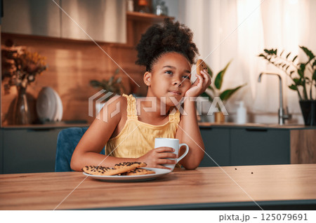 Eating cookies and holding cup. Cute black girl in casual clothes is at home at daytime 125079691