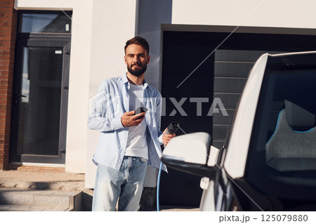 With smartphone. Young stylish man is with electric car at daytime With smartphone. Young stylish man is with electric car at daytime 125079880