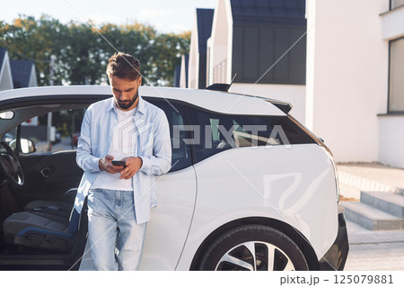 With smartphone. Young stylish man is with electric car at daytime 125079881