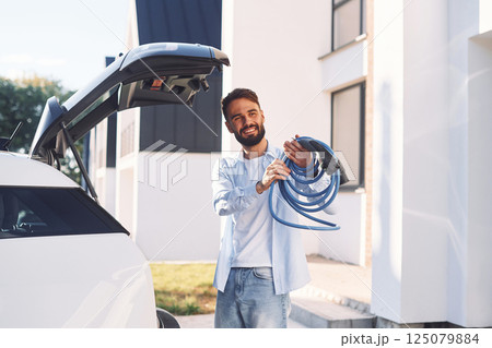Holding charging cable. Young stylish man is with electric car at daytime Holding charging cable. Young stylish man is with electric car at daytime 125079884