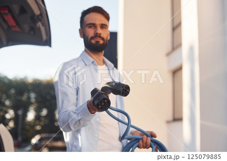 Holding charging cable. Young stylish man is with electric car at daytime Holding charging cable. Young stylish man is with electric car at daytime 125079885