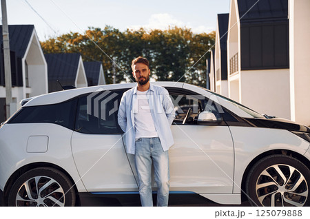 Standing and leaning on the vehicle. Posing for the camera. Young stylish man is with electric car at daytime 125079888