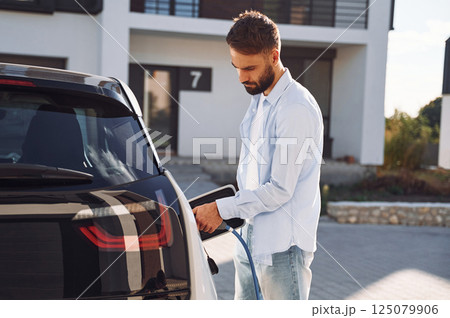 View from the side. Charging the vehicle. Young stylish man is with electric car at daytime 125079906