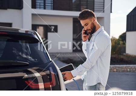 Charging the vehicle and using phone. Young stylish man is with electric car at daytime Charging the vehicle and using phone. Young stylish man is with electric car at daytime 125079918