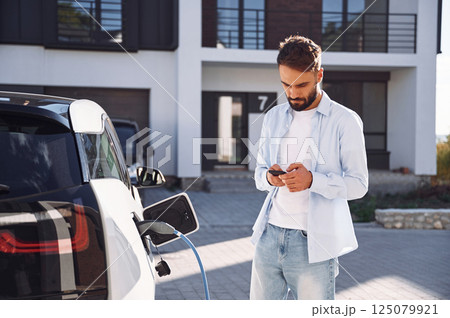 Waiting for charging complete, holding smartphone. Young stylish man is with electric car at daytime 125079921