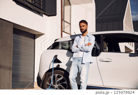 Standing with arms crossed and charging the vehicle. Young stylish man is with electric car at daytime 125079924