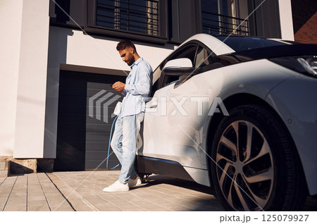 Sunny weather. Holding smartphone and charging the vehicle. Young stylish man is with electric car at daytime 125079927