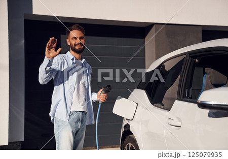 Hello, gesture. Holding charging wire. Young stylish man is with electric car at daytime Hello, gesture. Holding charging wire. Young stylish man is with electric car at daytime 125079935