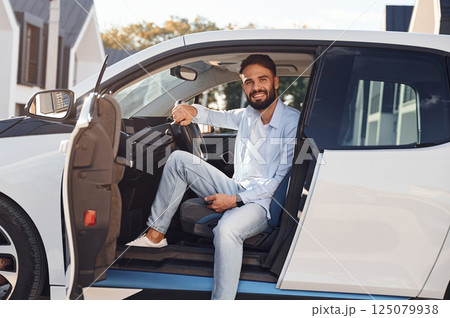 Sitting in the vehicle and smiling. Young stylish man is with electric car at daytime 125079938