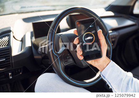 Process of charging is showing on smartphone. Close up view of man with his electric car 125079942