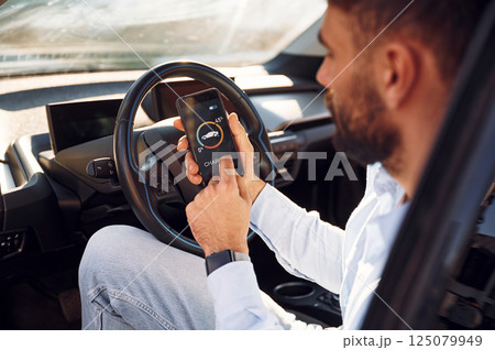 Monitoring charging procces by using smartphone. Young stylish man is with electric car at daytime Monitoring charging procces by using smartphone. Young stylish man is with electric car at daytime 125079949