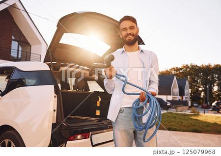 Against beautiful sunshine. Holding charging cables. Young stylish man is with electric car at daytime 125079968