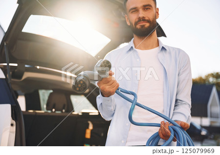 Against beautiful sunshine. Holding charging cables. Young stylish man is with electric car at daytime Against beautiful sunshine. Holding charging cables. Young stylish man is with electric car at daytime 125079969