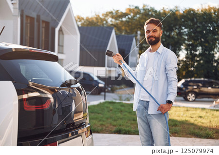 Conception of ecology. Standing with charging cable. Young stylish man is with electric car at daytime 125079974