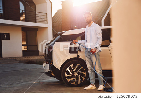Conception of ecology. Standing with charging cable. Young stylish man is with electric car at daytime 125079975