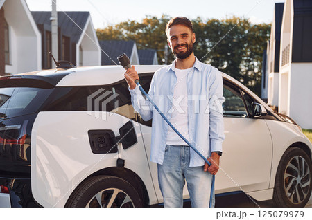 Conception of ecology. Standing with charging cable. Young stylish man is with electric car at daytime 125079979