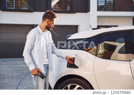 Plugging in charging cable. Young stylish man is with electric car at daytime Plugging in charging cable. Young stylish man is with electric car at daytime 125079980