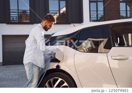 Plugging in charging cable. Young stylish man is with electric car at daytime 125079981