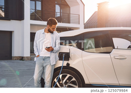 Charging the vehicle near the house. Holding smartphone. Young stylish man is with electric car at daytime Charging the vehicle near the house. Holding smartphone. Young stylish man is with electric car at daytime 125079984