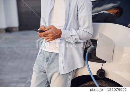 Standing with smartphone while automobile is charging. Close up view of man with his electric car 125079992