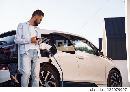 Standing and looking at vehicle that is being charging. Young stylish man is with electric car at daytime 125079997
