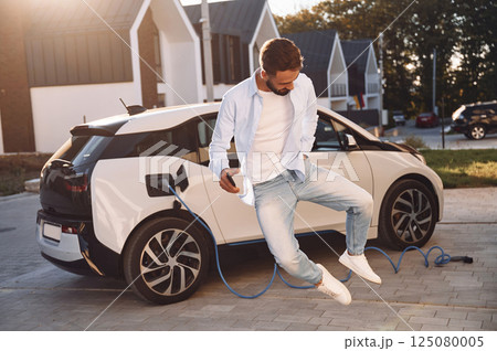 Jumping and having fun. Charging the vehicle. Young stylish man is with electric car at daytime Jumping and having fun. Charging the vehicle. Young stylish man is with electric car at daytime 125080005