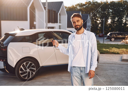 Standing and holding the keys. Young stylish man is with electric car at daytime Standing and holding the keys. Young stylish man is with electric car at daytime 125080011