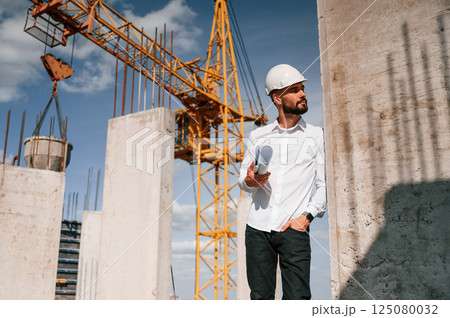 Standing and holding plan in hands. Man in uniform is working on the construction site 125080032