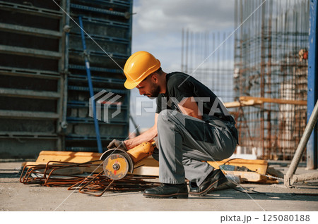 Sitting and cutting metal pieces by grinding machine. Man in uniform is working on the construction site 125080188