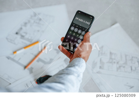 Calculating numbers by using smartphone. Plan is on the floor. The man is making repairs in the apartment 125080309