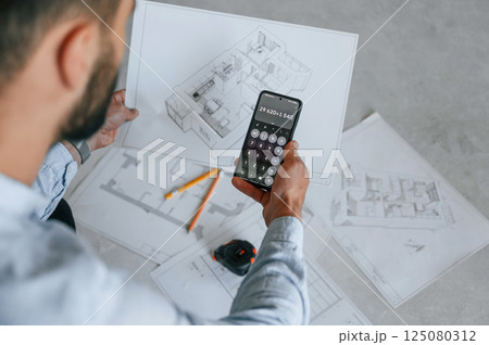 Calculating numbers by using smartphone. Plan is on the floor. The man is making repairs in the apartment Calculating numbers by using smartphone. Plan is on the floor. The man is making repairs in the apartment 125080312