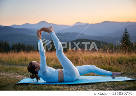 Woman practicing yoga outdoors in the mountains in a serene, natural setting. Female performing yoga pose on blue mat, with backdrop of beautiful mountain landscape at sunrise or sunset. Woman practicing yoga outdoors in the mountains in a serene, natural setting. Female performing yoga pose on blue mat, with backdrop of beautiful mountain landscape at sunrise or sunset. 125080770