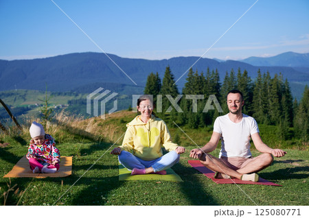 Young family doing yoga outdoors in scenic mountainous landscape in morning. Father, mother and daughter on yoga mats, with scenic view of mountains and forest in background under clear blue sky. Young family doing yoga outdoors in scenic mountainous landscape in morning. Father, mother and daughter on yoga mats, with scenic view of mountains and forest in background under clear blue sky. 125080771