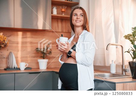 Standing on the kitchen with cup of drink. Beautiful pregnant woman is indoors at home 125080833