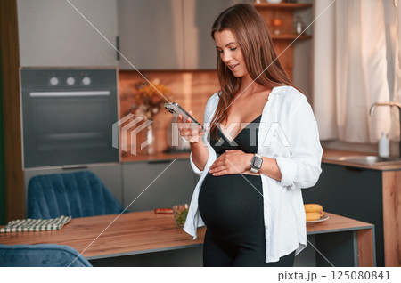 Standing with smartphone in hand. Beautiful pregnant woman is indoors at home Standing with smartphone in hand. Beautiful pregnant woman is indoors at home 125080841